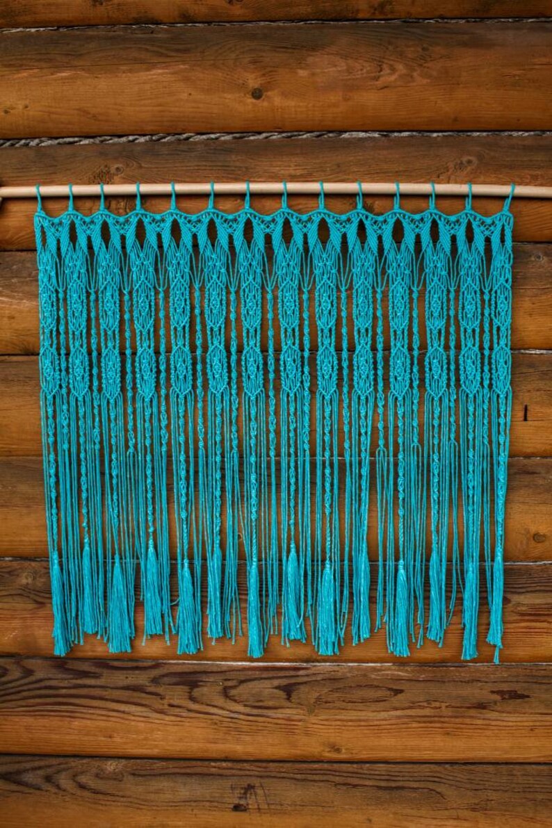 May include: A turquoise macrame wall hanging with a wooden dowel rod at the top. The macrame has a fringe at the bottom and is made with a knot pattern.