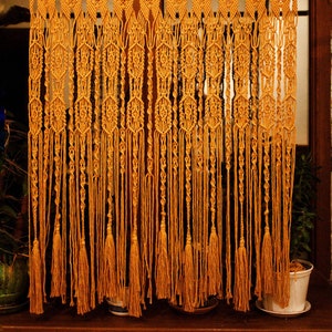 May include: A macrame curtain with a geometric pattern and tassels. The curtain is made of natural fiber rope and is a warm, golden yellow color.
