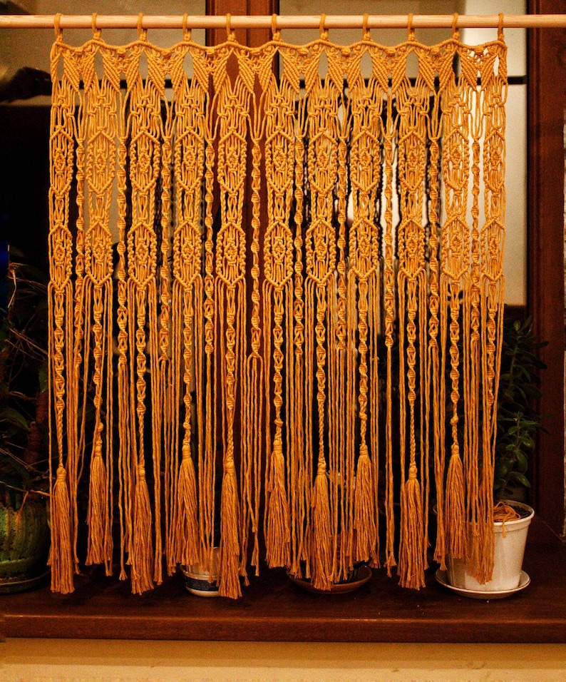 May include: A macrame curtain with a geometric pattern and tassels. The curtain is made of natural fiber rope and is a warm, golden yellow color.