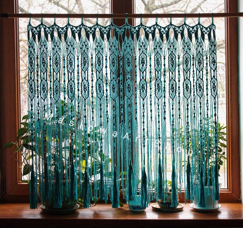 May include: Teal macrame curtain with a geometric pattern, hanging in a window with green plants in pots on either side.