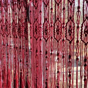 May include: A pink macrame curtain with a geometric pattern hangs in a window. The curtain is made of thick yarn and has a fringe at the bottom.