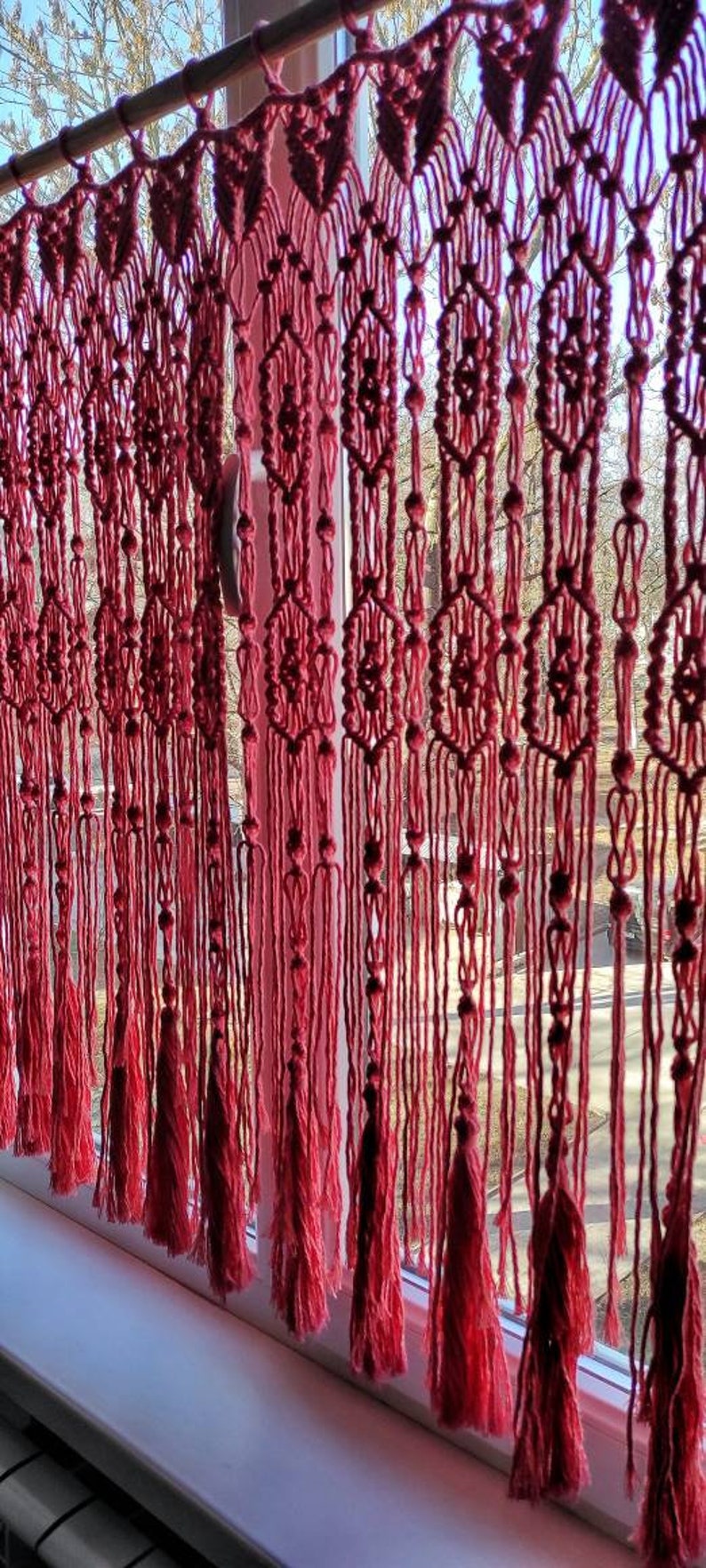 May include: A pink macrame curtain with a geometric pattern hangs in a window. The curtain is made of thick yarn and has a fringe at the bottom.