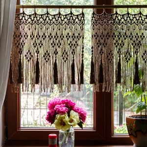 May include: Two macrame curtains with tassels hang in a window with a bouquet of pink and white peonies in a glass vase on the windowsill.