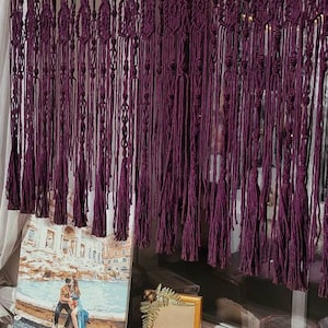 May include: A purple macrame wall hanging with tassels. The hanging is made of thick cord and has a woven pattern. It is hanging on a wooden rod.