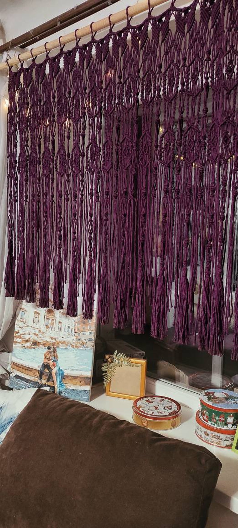 May include: A purple macrame wall hanging with tassels. The hanging is made of thick cord and has a woven pattern. It is hanging on a wooden rod.