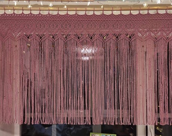 May include: A pink macrame curtain with a decorative top border and beaded accents. The curtain is hanging from a wooden rod.