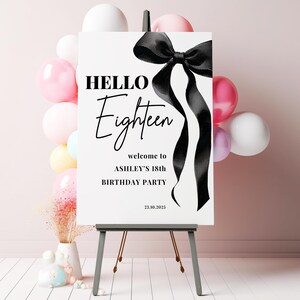May include: A white welcome sign with black text and a black satin ribbon bow. The text reads "HELLO Eighteen welcome to ASHLEY'S 18th BIRTHDAY PARTY 23.10.2023".