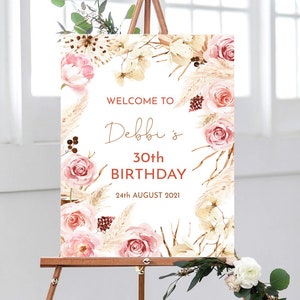 May include: Welcome to Debbi's 30th Birthday sign featuring a floral design with pink roses, pampas grass, and greenery. The sign is on a white background and has a rustic feel.