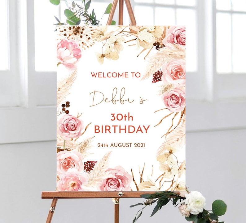May include: Welcome to Debbi's 30th Birthday sign featuring a floral design with pink roses, pampas grass, and greenery. The sign is on a white background and has a rustic feel.