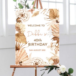 May include: Welcome to Debbi's 40th Birthday party sign with a white background and a floral design featuring dried pampas grass, leaves, and flowers in shades of brown and beige.