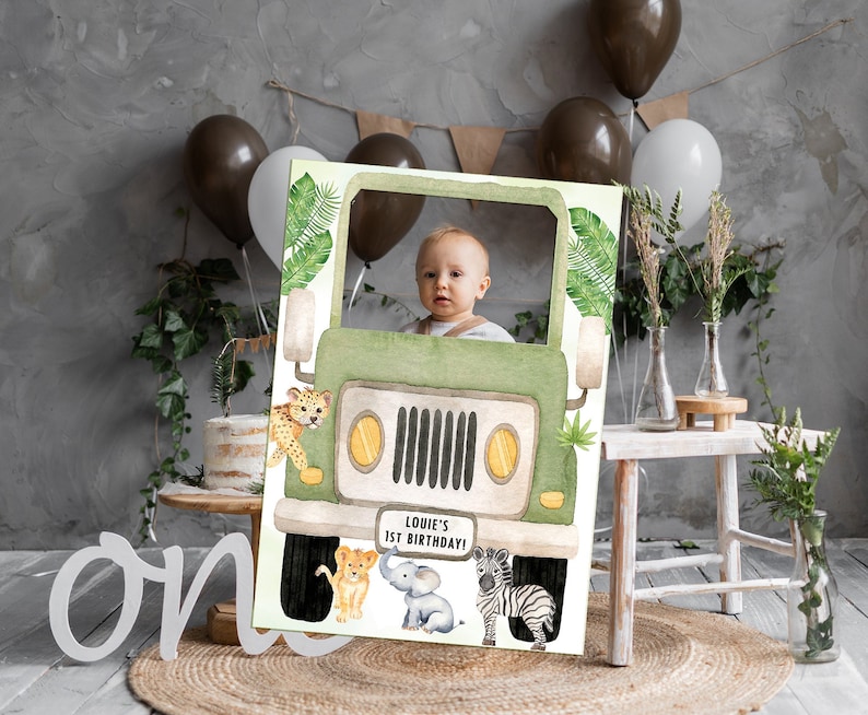 May include: A green jeep photo booth prop with a cut-out for a face. The jeep has a "Louie's 1st Birthday!" sign on the front. There are animal illustrations around the jeep, including a lion, elephant, and zebra.