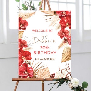 May include: Welcome to Debbi's 30th Birthday sign with a tropical theme. The sign features red orchids, dried palm leaves, and a white background. The text is in a brown font.