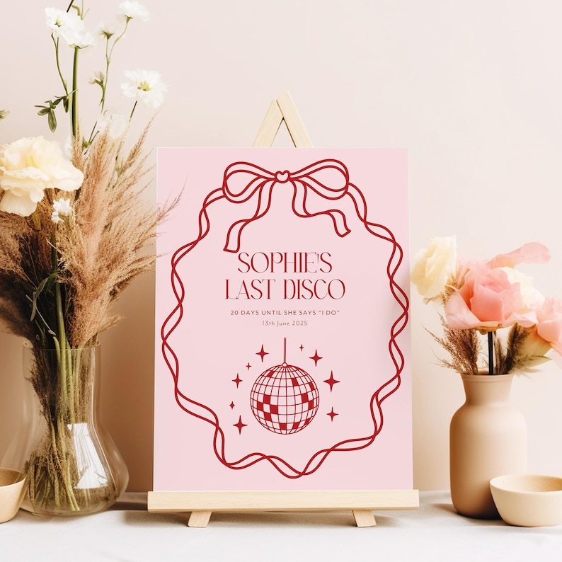 May include: A pink sign with a red border and a red bow at the top. The sign says "Sophie's Last Disco" and "20 days until she says 'I do' 13th June 2023". There is a red disco ball in the center of the sign.