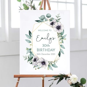 May include: Welcome to Emily's 30th Birthday sign with a gold geometric frame and white flowers with green leaves.