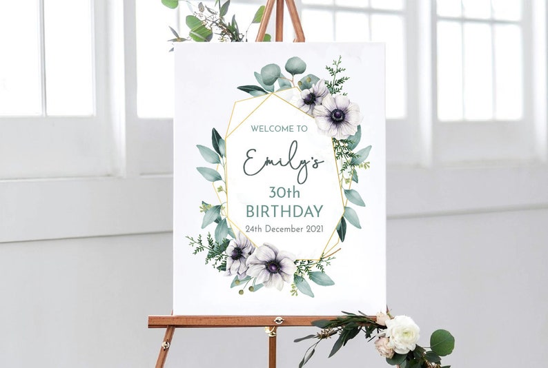May include: Welcome to Emily's 30th Birthday sign with a gold geometric frame and white flowers with green leaves.