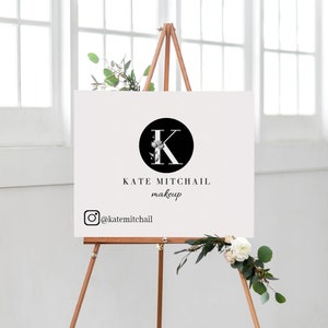 May include: A white sign with a black circle containing a white letter K with floral accents. The text "KATE MITCHAIL makeup" is below the circle. The Instagram logo and handle "@katemitchail" are below the text. The sign is on a wooden easel.