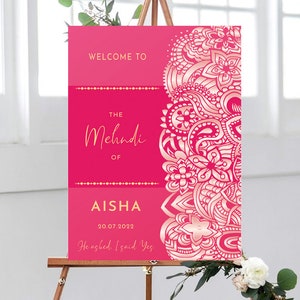 May include: A pink welcome sign with gold lettering announcing "The Mehndi of Aisha" with the date "20.07.2022" and the text "He asked. I said Yes."  The sign features a decorative pink and gold mandala design.
