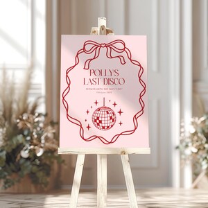 May include: A pink sign with a red border and a red bow at the top. The sign says "Polly's Last Disco" and "20 days until she says 'I do'" with the date "13th June 2023". A red disco ball is in the center of the sign.
