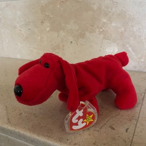 May include: A plush, red dog toy with a black nose and black eyes. The toy has long, floppy ears and a small tail. A clear tag with the text "Ty Beanie Babies" is attached to the toy.