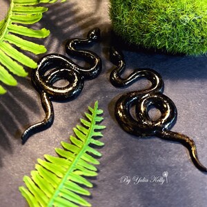 Könnte beinhalten: Zwei schwarze Schlangenohrringe mit glänzender Oberfläche. Die Ohrringe liegen auf einem grauen Hintergrund mit grünem Laub.