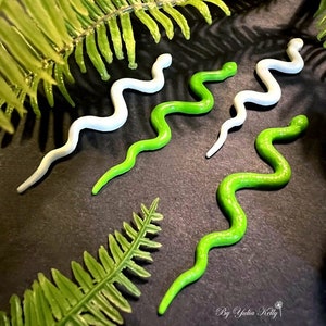 May include: Three decorative snake-shaped polymer clay earrings. Two are green and one is white. The earrings are displayed on a black surface with green fern leaves.