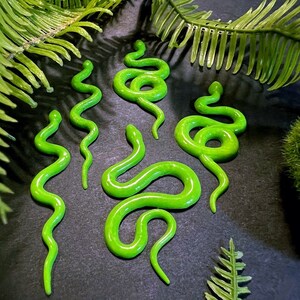May include: Five green snake-shaped resin earrings on a black background with green fern leaves. By Yulia Kelly.