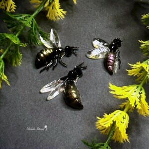 Può includere: Tre figurine di api in resina con ali trasparenti. Le api sono nere con accenti dorati e hanno un bagliore iridescente viola. Le api sono disposte su una superficie grigia con fiori gialli.