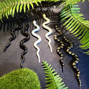 Op de afbeelding: Vijf paar slangen oorringen in zwart, wit en zwart en goud. De oorringen zijn gemaakt van polymer clay en zijn gevormd als slangen. De oorringen worden getoond op een donkergrijze achtergrond met groene varenbladeren.