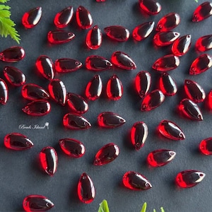 May include: A large quantity of red teardrop-shaped glass beads. The beads are scattered on a black surface.