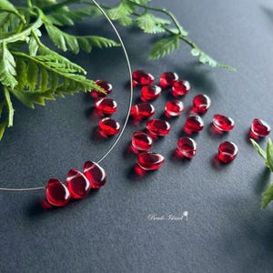 May include: A scattering of red glass teardrop beads on a dark gray surface. Three of the beads are strung on a silver wire.
