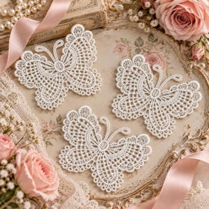 Può includere: Tre applicazioni a farfalla in pizzo bianco con intricati motivi traforati e bordi smerlati. Ogni farfalla presenta ali simmetriche con dettagliate cuciture a rete e due sottili antenne, presentate come singoli pezzi decorativi.