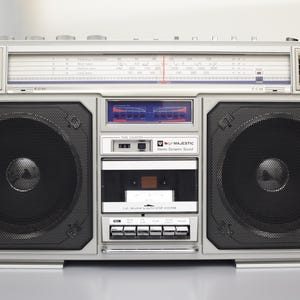 May include: A silver and black cassette boombox with a silver faceplate and two large speakers. The boombox has a cassette player, a radio tuner, and a volume knob. The brand name "Majestic" is printed on the front of the boombox.