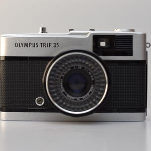Op de afbeelding: Een zilveren Olympus Trip 35 filmcamera met een zwarte lens en een zwart, getextureerd lichaam. De camera heeft een bandje.