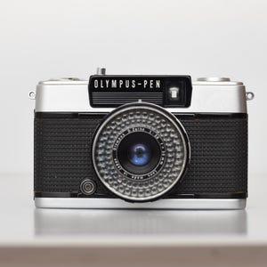Puede incluir: Una cámara de película Olympus-Pen 35 mm de color plateado y negro con una lente negra y un cuerpo negro texturizado.