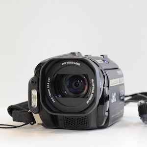 Puede incluir: Una videocámara JVC Everio negra con un objetivo JVC Video Lens con zoom óptico de 10x, flash integrado y una correa de transporte de tela negra. El barril del objetivo muestra especificaciones técnicas, incluyendo 3,2-32mm y 1:1,8.
