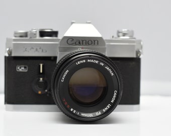 Może przedstawiać: Srebrny aparat fotograficzny Canon AE-1 Program z obiektywem Canon FD 100mm 1:2.8 ssc. Obiektyw jest czarny i ma nadrukowany tekst "LENS MADE IN JAPAN CANON LENS FD 100mm 1:2.8 ssc".