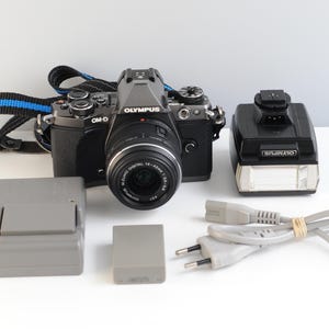 Puede incluir: Una cámara digital Olympus OM-D negra con objetivo, una correa negra y azul, un flash externo negro, un cargador de batería gris, una batería gris y un cable de alimentación gris con un enchufe de dos clavijas.