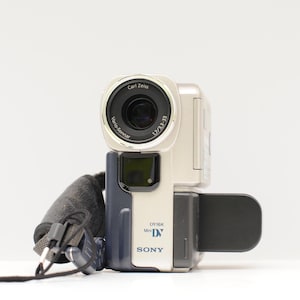 Może przedstawiać: Srebrno-granatowa kamera cyfrowa Sony MiniDV z obiektywem Carl Zeiss Vario-Sonnar. Urządzenie posiada czarny pasek na rękę z małym pilotem, boczny rozkładany ekran oraz wyraźnie widoczne oznaczenia marki i modelu na obudowie.
