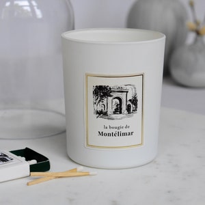 Puede incluir: Vela blanca con una etiqueta en blanco y negro que dice "la bougie de Montélimar". La etiqueta presenta un dibujo de un arco con árboles a cada lado. La vela está sobre una superficie de mármol blanco con una caja de cerillas.