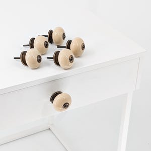 May include: Six cream-colored ceramic drawer knobs with a brown metal accent in the center. The knobs are arranged on a white dresser with a drawer.
