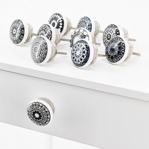 Könnte beinhalten: Ein Satz von zehn weißen Keramik-Schubladengriffen mit schwarzen und grauen Mandala-Designs. Die Griffe haben silberne Metallschrauben.