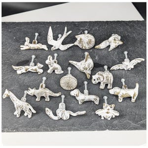 Op de afbeelding: Een verzameling van veertien decoratieve kastknoppen in diverse diervormen, waaronder een giraf, dinosaurus, slang, bij, hond, vos, konijn, vogel en vleermuis, afgewerkt met een verweerde witte en gouden verflaag en metalen stelen.