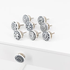Peut inclure: Un ensemble de neuf boutons de placard ronds ornés de motifs complexes noirs et blancs. Chaque bouton présente une face circulaire avec des dessins géométriques ou floraux détaillés et une base en métal argenté avec une vis filetée.