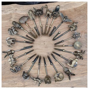 Może przedstawiać: Kolekcja dekoracyjnych metalowych podwiązek do zasłon z motywami zwierzęcymi, w tym lis, żyrafa, wiewiórka i nietoperz. Podwiązki ułożone są w okrąg na drewnianej powierzchni, prezentując szczegółowe wzory i antyczne wykończenie.