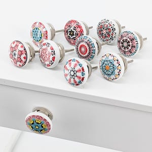Op de afbeelding: Set van 10 keramische kastknoppen met kleurrijke mandala-ontwerpen. De knoppen zijn wit met roze, rode, blauwe en gele accenten. Elke knop heeft een zilveren schroef.