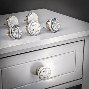 Puede incluir: Una colección de pomos de armario blancos y plateados con un diseño vintage. Los pomos presentan texto e ilustraciones en negro, incluyendo las palabras "Olive Oil Soft Soap" y "Foresters England". Los pomos se muestran en un armario gris.