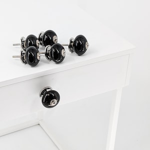 Könnte beinhalten: Sechs schwarze Keramik-Schrankknöpfe mit silbernen Akzenten auf einer weißen Oberfläche. Die Knöpfe sind rund und haben eine glatte Oberfläche.