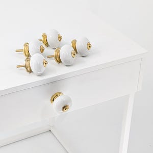Könnte beinhalten: Eine Sammlung weißer Keramik-Schrankknöpfe mit goldfarbener Hardware. Die Knöpfe sind oval geformt und haben eine glänzende Oberfläche. Ein Knopf ist an einer weißen Schublade befestigt, die anderen liegen auf einer weißen Oberfläche.