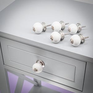 Peut inclure: Ensemble de six boutons de meuble en céramique blanche avec des bases et tiges en métal argenté. Un bouton est fixé à un tiroir gris, les cinq autres sont disposés sur la surface grise. Les boutons sont ronds.