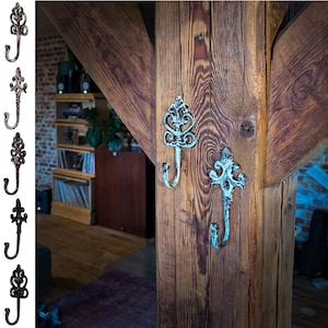 Könnte beinhalten: Zwei dekorative Metallhaken mit verzierten Designs sind an einem Holzbalken montiert. Die Haken sind in einem abgewetzten Weiß lackiert und haben einen rustikalen Look.